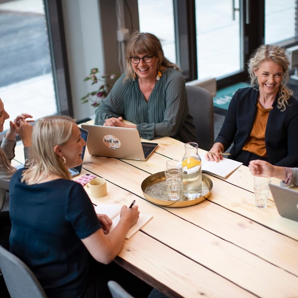 Four people sitting around a wooden table in a meeting room, two women smiling and talking, laptops with FAB HR stickers, glass jug with water and lemon on a tray